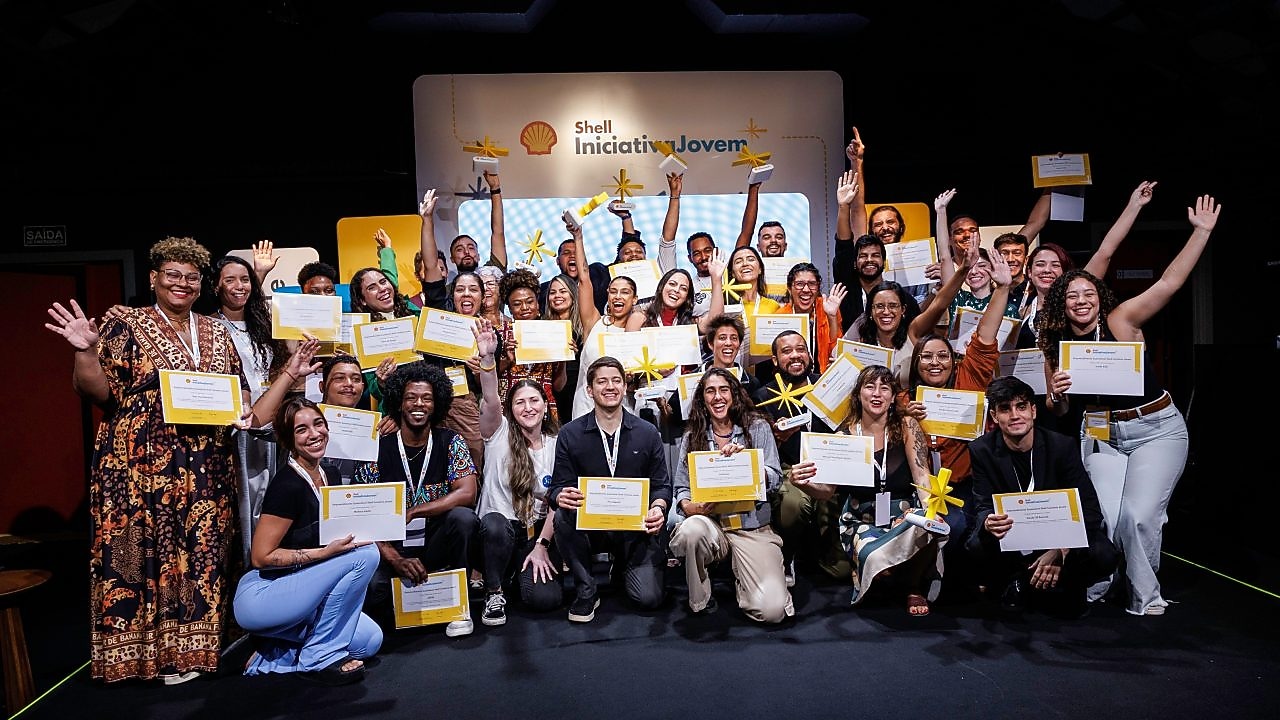Grupo diverso de jovens empreendedores posando sorridentes com certificados nas mãos em evento de formatura do programa Shell Iniciativa Jovem. Ao fundo, painel com o logotipo da Shell e do programa. Todos celebram com entusiasmo, alguns segurando troféus em formato de estrela amarela.