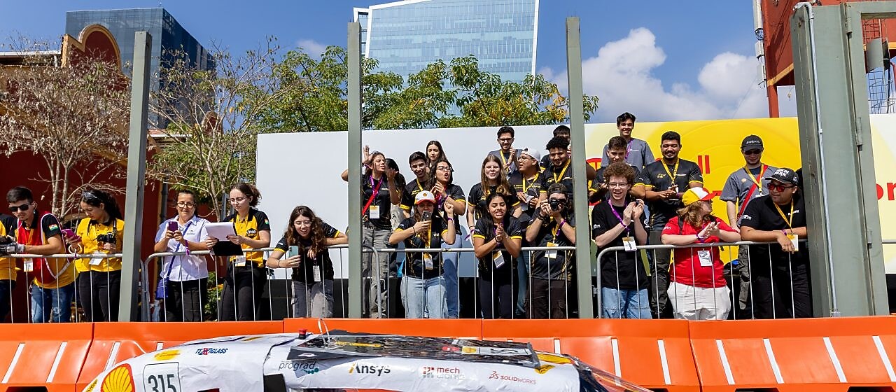 Protótipo de carro preto e aerodinâmico, número 315, decorado com logotipos de patrocinadores como Shell, Ansys e SolidWorks, passando em frente a uma arquibancada. Um grupo de pessoas, em sua maioria jovens com crachás, aplaude e fotografa o veículo. Ao fundo, prédios modernos e árvores completam o cenário ensolarado.