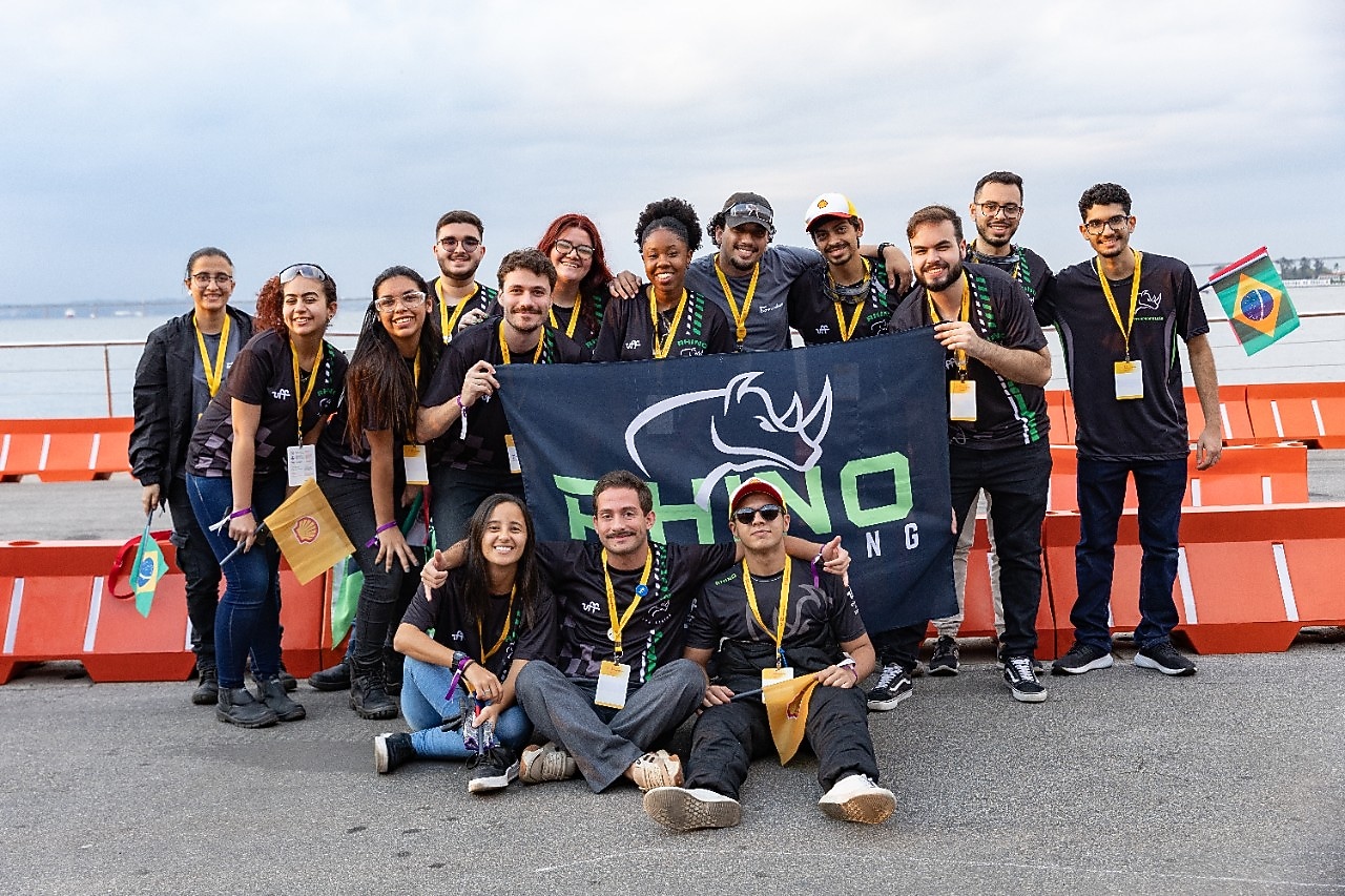 Grupo de jovens sorridentes posando juntos ao ar livre, usando camisetas pretas da equipe Rhino Racing e crachás amarelos. Dois membros estão sentados à frente, segurando uma bandeira preta com o logotipo de um rinoceronte e o nome Rhino Racing. Ao fundo, há barreiras laranjas e o mar. Algumas pessoas seguram bandeiras do Brasil e da Shell.