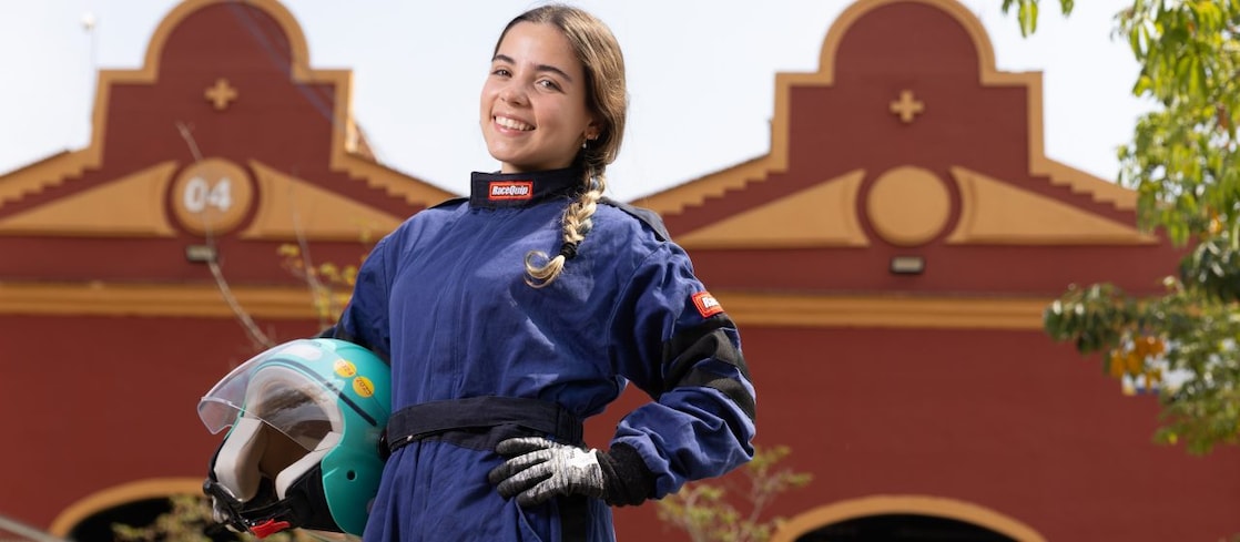 Menina sorridente vestida com macacão de corrida azul, luvas brancas e pretas, segurando um capacete verde com viseira transparente. Ela está ao ar livre, em frente a um prédio vermelho com detalhes em amarelo e o número 04 no topo.
