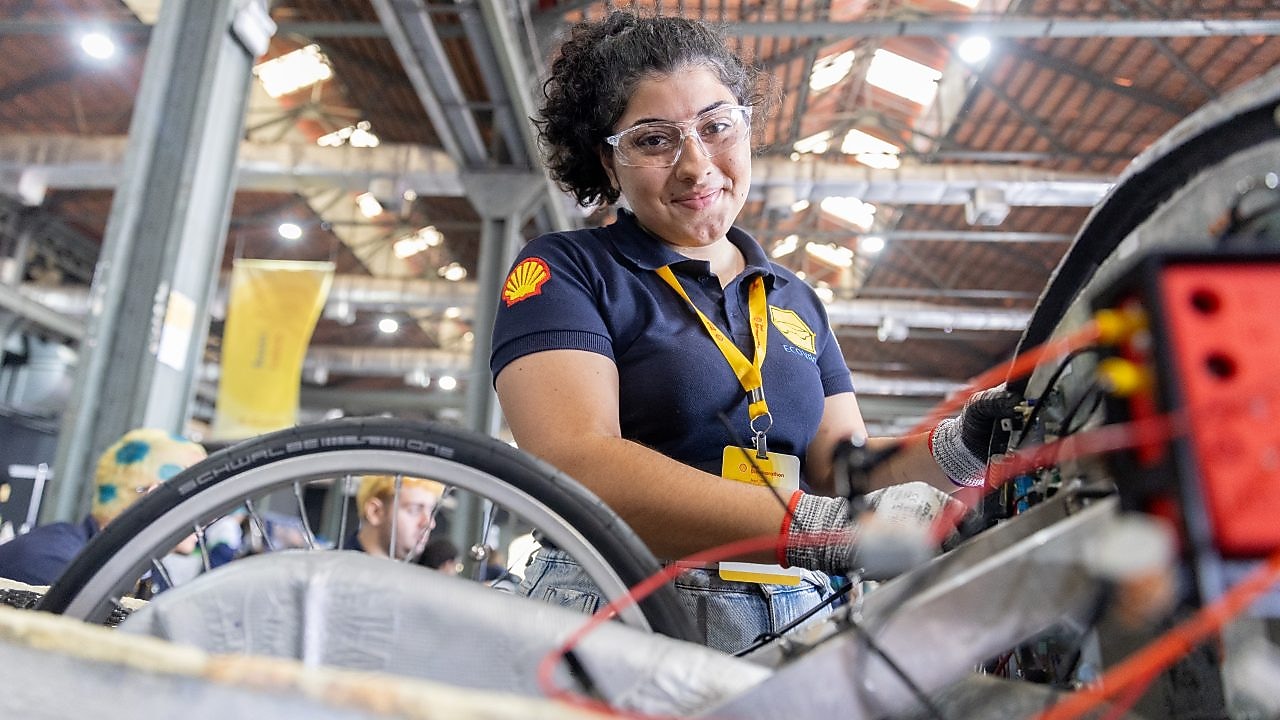 Menina de óculos de proteção e luvas de trabalho, usando camiseta azul-marinho com logotipo da Shell e crachá amarelo, sorri enquanto trabalha na montagem de um veículo em ambiente industrial.