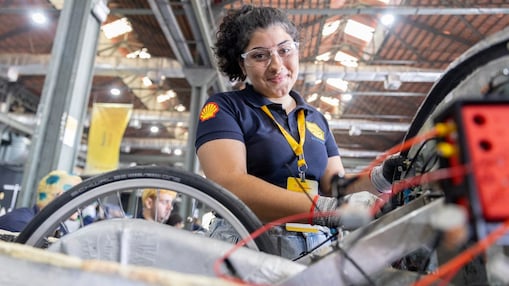 Menina de óculos de proteção e luvas de trabalho, usando camiseta azul-marinho com logotipo da Shell e crachá amarelo, sorri enquanto trabalha na montagem de um veículo em ambiente industrial.