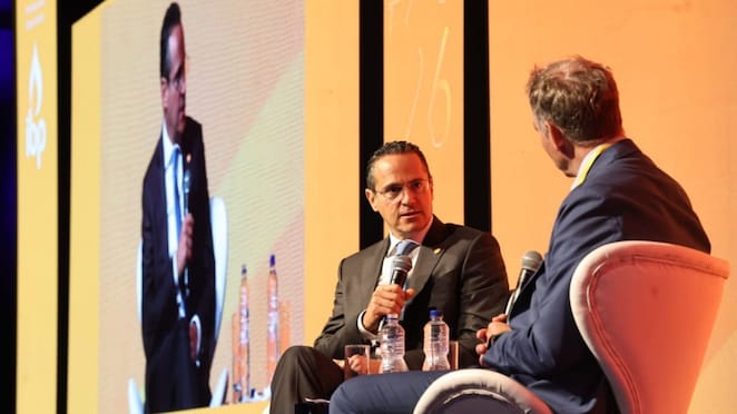 Dois executivos participam de uma conversa no palco durante um evento, sentados em poltronas e segurando microfones. Ao fundo, um telão exibe a imagem de um dos participantes.