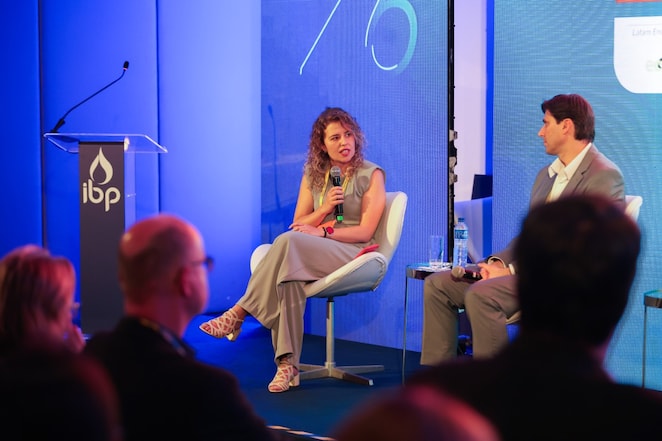 Mulher fala ao microfone durante um painel em um evento, sentada ao lado de outro participante. Ao fundo, grandes telas azuis compõem o palco, que também conta com um púlpito com o logotipo do IBP. Na parte inferior da imagem, aparecem pessoas da plateia.
