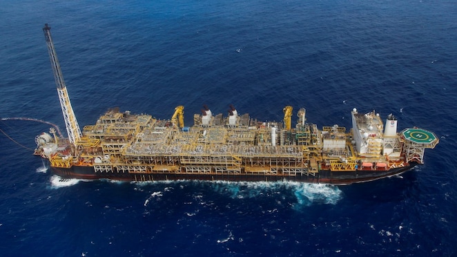 Plataforma de petróleo offshore em alto-mar, vista aérea, com estrutura industrial extensa e heliponto à direita, cercada por águas profundas do oceano.