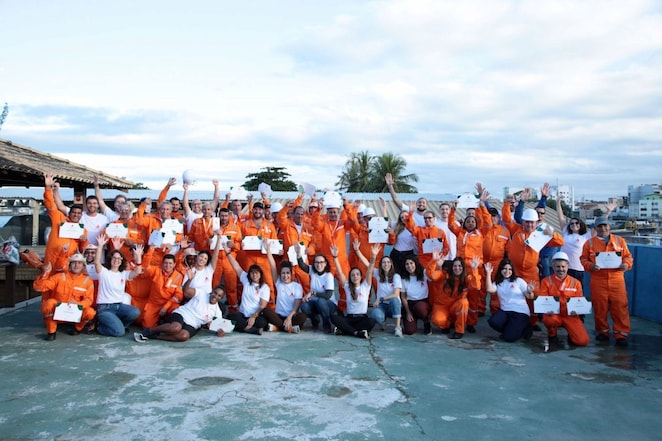 Grupo grande de aproximadamente 50 pessoas reunidas ao ar livre em uma área industrial. A maioria veste macacões laranja de segurança e alguns usam capacetes brancos, enquanto outros estão com camisetas brancas. Muitas pessoas seguram certificados e acenam para a câmera com os braços erguidos, em clima de celebração. Ao fundo, há estruturas industriais, telhados, algumas palmeiras e prédios urbanos sob um céu nublado. A foto transmite um momento de reconhecimento coletivo e conclusão de uma atividade ou treinamento.