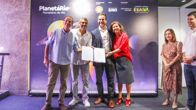 Grupo de pessoas em pé, sorrindo, durante cerimônia no Planetário do Rio. Quatro pessoas ao centro seguram um documento aberto, diante de um painel com a identidade visual do Planetário do Rio e logos de patrocinadores. Ao fundo, ilustrações de planetas. Outras pessoas acompanham a cena nas laterais do palco.