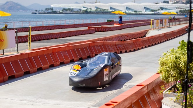 Veículo preto aerodinâmico com canopy transparente e número 324, participando da Shell Eco-marathon em pista ao ar livre, com barreiras laranjas de segurança, água e prédios modernos ao fundo em dia ensolarado.