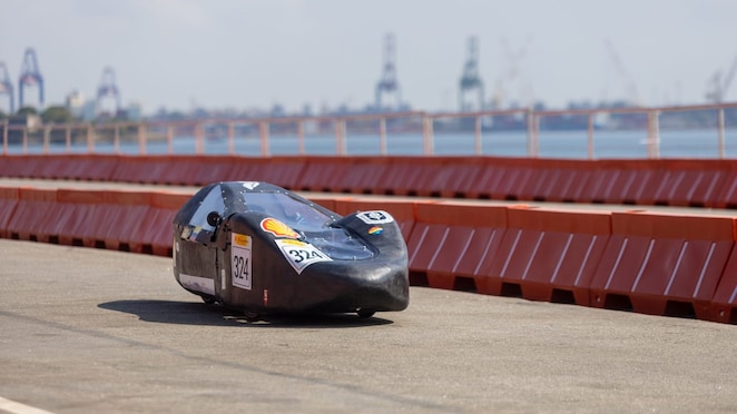 Veículo protótipo preto e aerodinâmico, com canopy transparente e logotipo da Shell, circula em pista asfaltada ao lado da água. Marcado com o número 324 e vários adesivos de patrocinadores, participa de uma competição de eficiência energética próximo a guindastes e barreiras vermelhas de segurança.