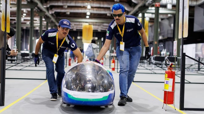 Dois integrantes de uma equipe universitária empurram um veículo de alta eficiência energética em uma área de competição da Shell Eco-marathon. O carro tem formato compacto e aerodinâmico, com cabine transparente e detalhes em azul, verde e branco. Os participantes vestem uniformes azul-marinho e credenciais do evento.