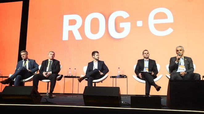 Cinco homens participam de um painel em um palco, sentados em poltronas brancas. Um dos participantes fala ao microfone, enquanto os demais acompanham a conversa. Ao fundo, um grande painel em tons de laranja exibe o logotipo “ROG-e”.