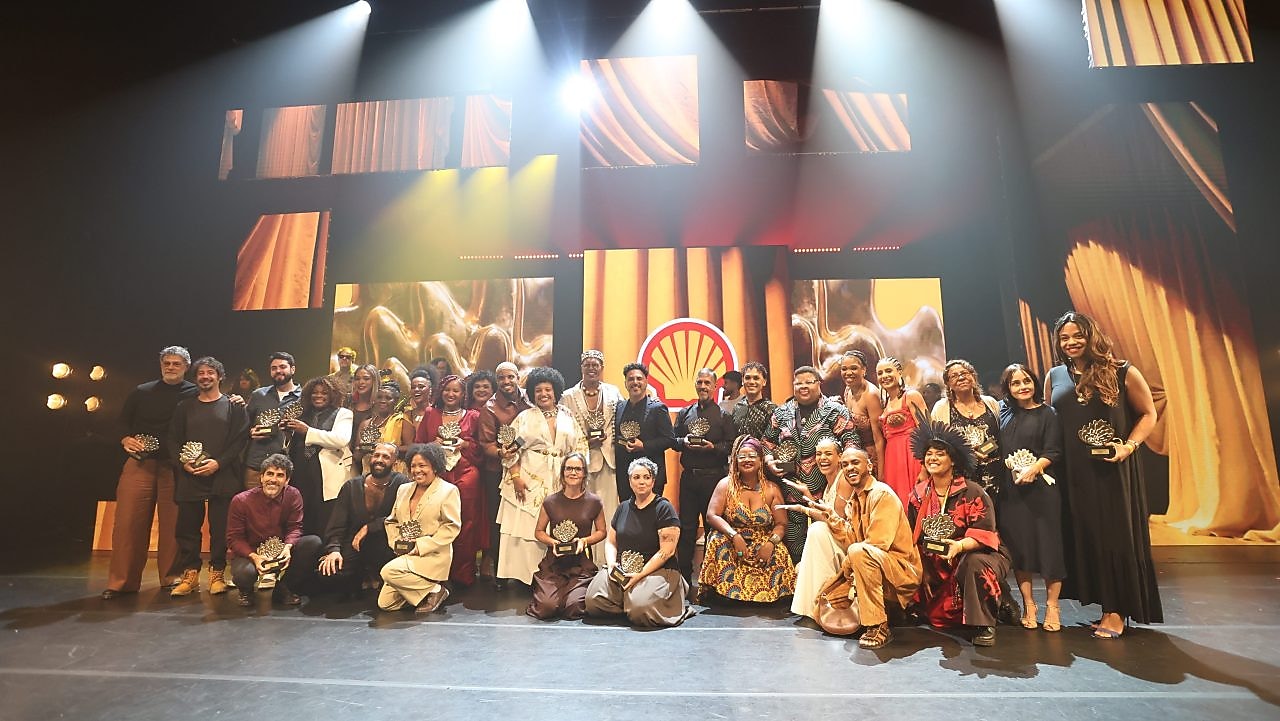 Grupo de premiados reunidos no palco durante uma cerimônia do Prêmio Shell de Teatro. Todos seguram troféus e posam para a foto em duas fileiras. Ao fundo, painéis iluminados em tons dourados e o logotipo da Shell. A iluminação de palco destaca o ambiente e reforça o clima de celebração.