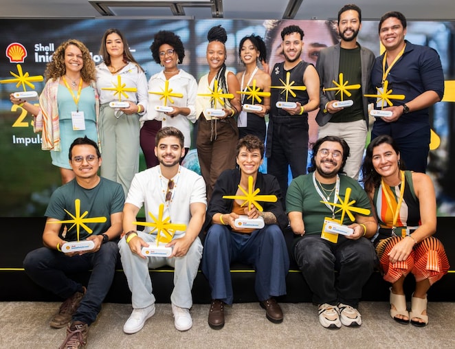 Grupo diverso de 14 pessoas posa para foto em um palco, segurando troféus amarelos em formato de estrela. Elas estão organizadas em duas fileiras, algumas em pé e outras sentadas, sorrindo para a câmera. Ao fundo, há um painel com a marca Shell Iniciativa Jovem. O ambiente é interno, com iluminação profissional, e todas usam crachás de identificação, indicando um evento corporativo ou de premiação.
