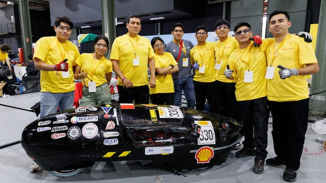 Grupo de participantes da Shell Eco-marathon posa ao lado de um veículo de eficiência energética preto, identificado com o número 330 e o logotipo da Shell. A equipe, formada por dez pessoas, veste majoritariamente camisetas amarelas e equipamentos de segurança. A foto foi registrada em uma área interna de competição, com boxes e estruturas técnicas ao fundo.