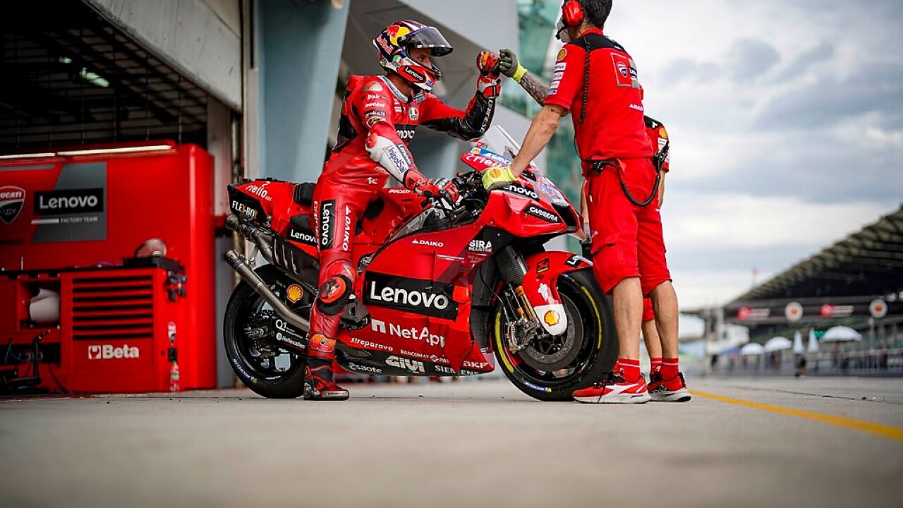 Piloto da equipe Ducati Corse saindo de motocicleta do box e cumprimentando equipe