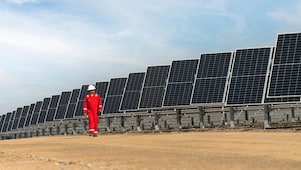 Em primeiro plano, placas solares de usina solar que abastecem instalação industrial. Ao fundo, instalação industrial sob céu azulado com algumas nuvens.
