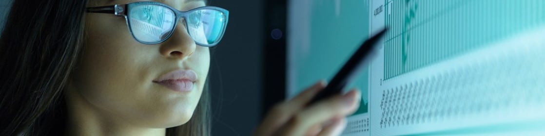 Jovem mulher de óculos aponta para uma tela exibindo alguns gráficos