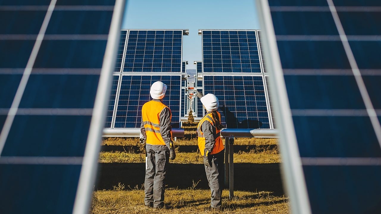 Campo de painéis de energia solar fotovoltaica onde se vê um homem e uma mulher mulher paramentados trabalhando