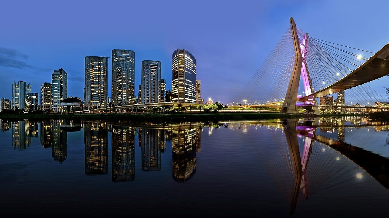 O horizonte de São Paulo em uma vista panorâmica no entardecer, mostrando uma concentração de prédios comerciais e a icônica ponte estaiada 'Ponte Octavio Frias de Oliveira' 