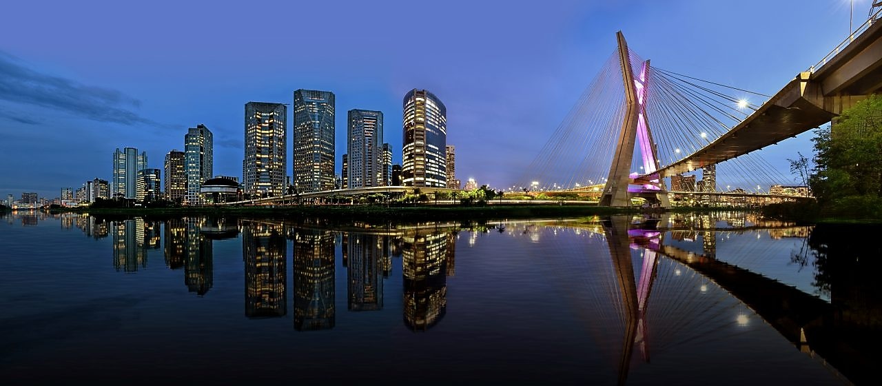 O horizonte de São Paulo em uma vista panorâmica no entardecer, mostrando uma concentração de prédios comerciais e a icônica ponte estaiada 'Ponte Octavio Frias de Oliveira'