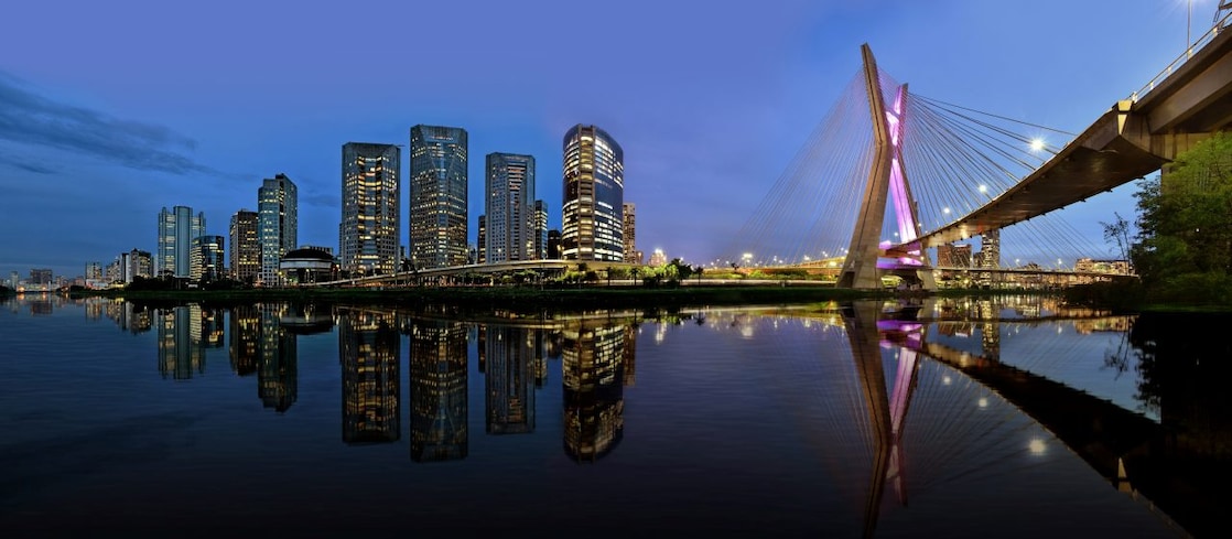 O horizonte de São Paulo em uma vista panorâmica no entardecer, mostrando uma concentração de prédios comerciais e a icônica ponte estaiada 'Ponte Octavio Frias de Oliveira'