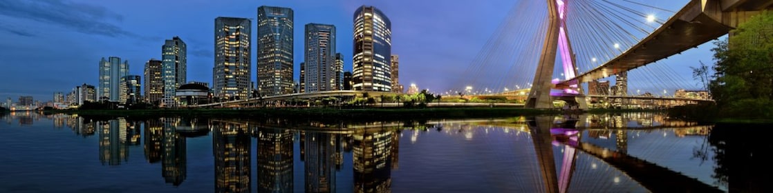 O horizonte de São Paulo em uma vista panorâmica no entardecer, mostrando uma concentração de prédios comerciais e a icônica ponte estaiada 'Ponte Octavio Frias de Oliveira'