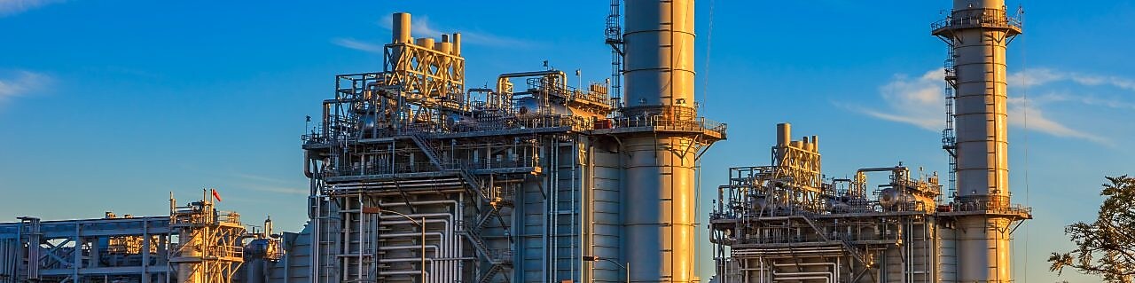 Na imagem, temos uma vista de uma planta para extração de gás natural, com uma pequena vegetação na frente da construção.