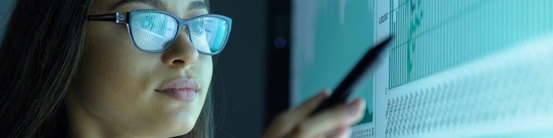 Jovem mulher de óculos aponta para uma tela exibindo alguns gráficos