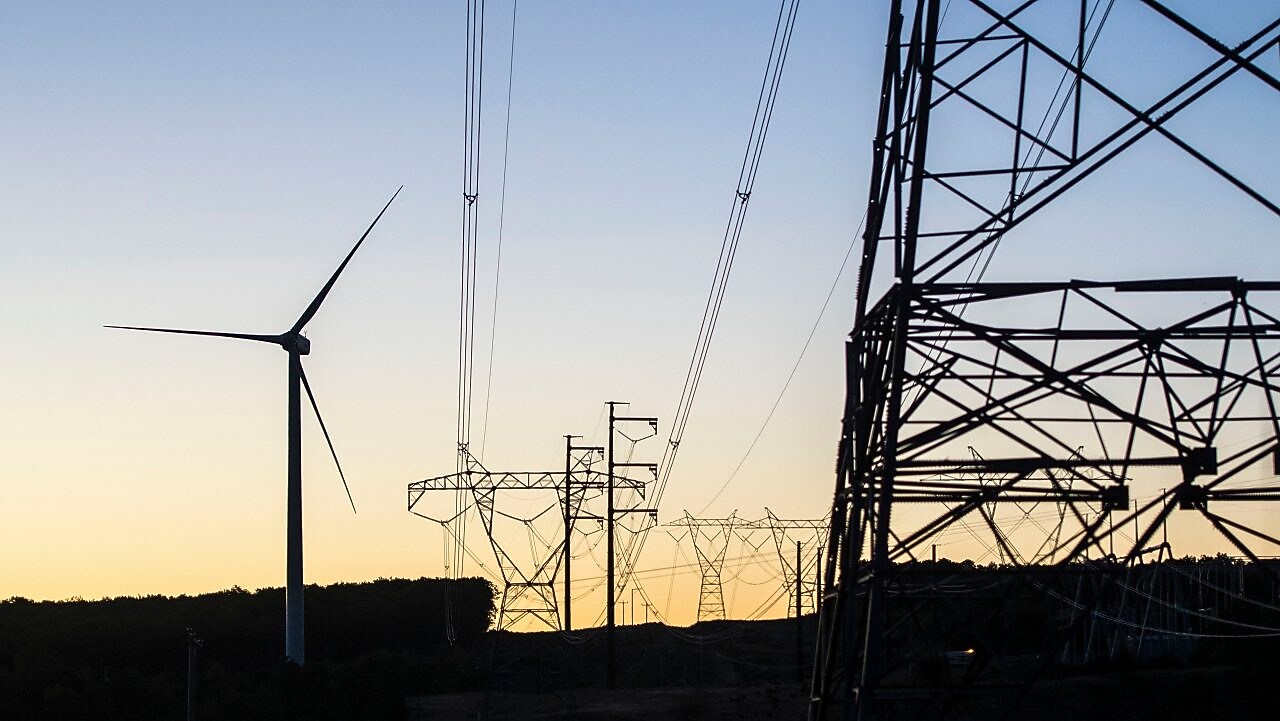 Vista de um campo com turbinas eólicas e cabos de transmissão ao fim da tarde.