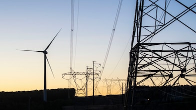 Vista de um campo com turbinas eólicas e cabos de transmissão ao fim da tarde.
