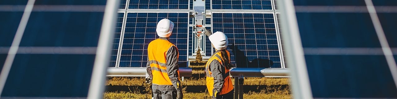 Campo de painéis de energia solar fotovoltaica onde se vê um homem e uma mulher mulher paramentados trabalhando