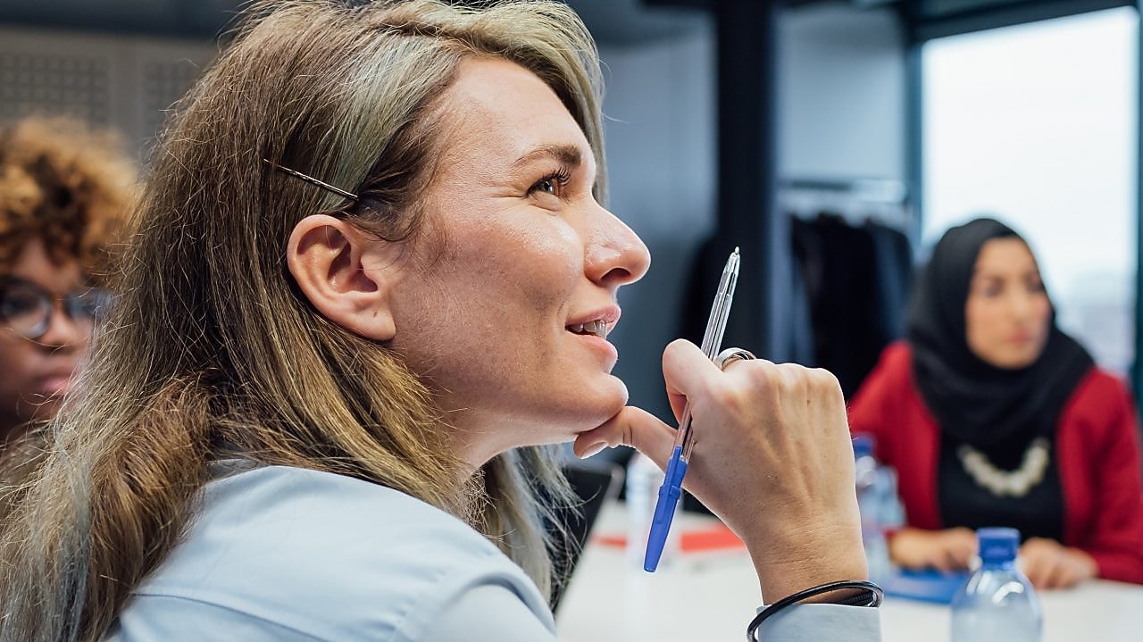 três mulheres de traje social atentas em uma mesa de reunião.