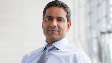 Homem de cabelo escuro, vestindo traje social, senta em uma cadeira olhando pra câmera