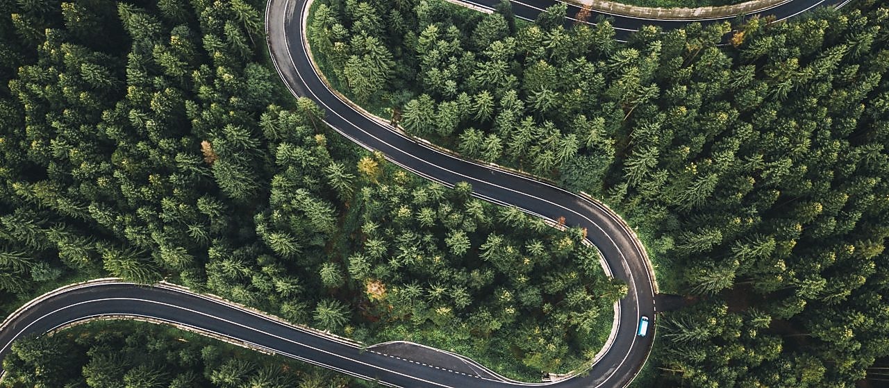 Vista aérea de uma estrada sinuosa cercada por uma densa floresta de pinheiros. A estrada faz curvas acentuadas em forma de ‘S’, cortando o verde intenso das árvores. Um carro azul percorre o trecho, destacando-se no cenário natural.