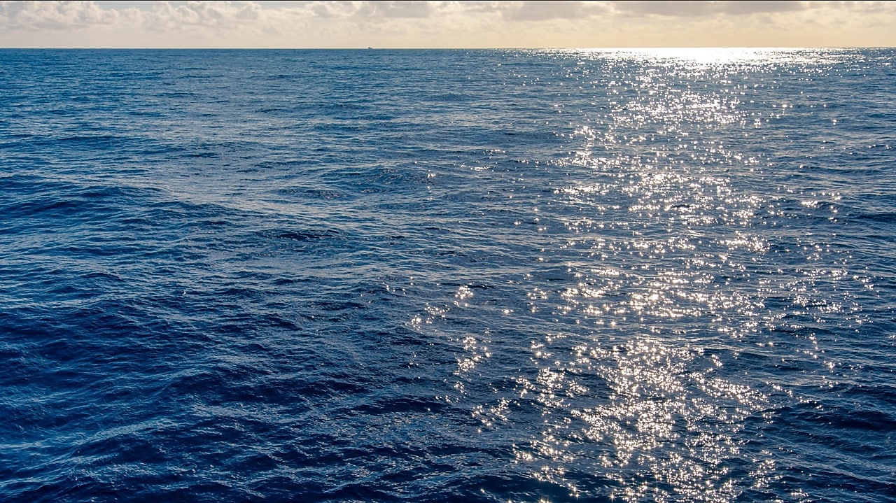 Superfície do oceano iluminada pelo sol, com pequenas ondas e reflexos dourados se espalhando pela água azul. O horizonte mostra o encontro do mar com o céu claro e algumas nuvens dispersas.