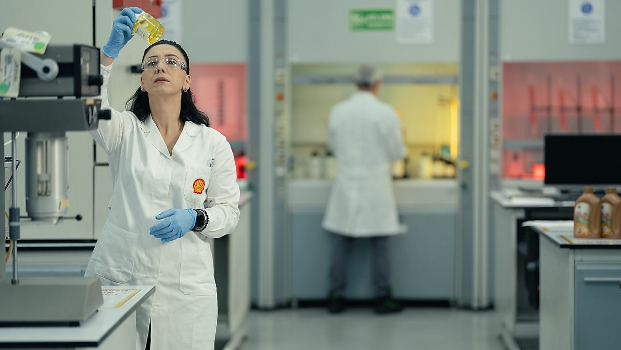 Cientista em laboratório da Shell usando jaleco, óculos de proteção e luvas, segurando um frasco com líquido amarelo enquanto analisa sua composição.