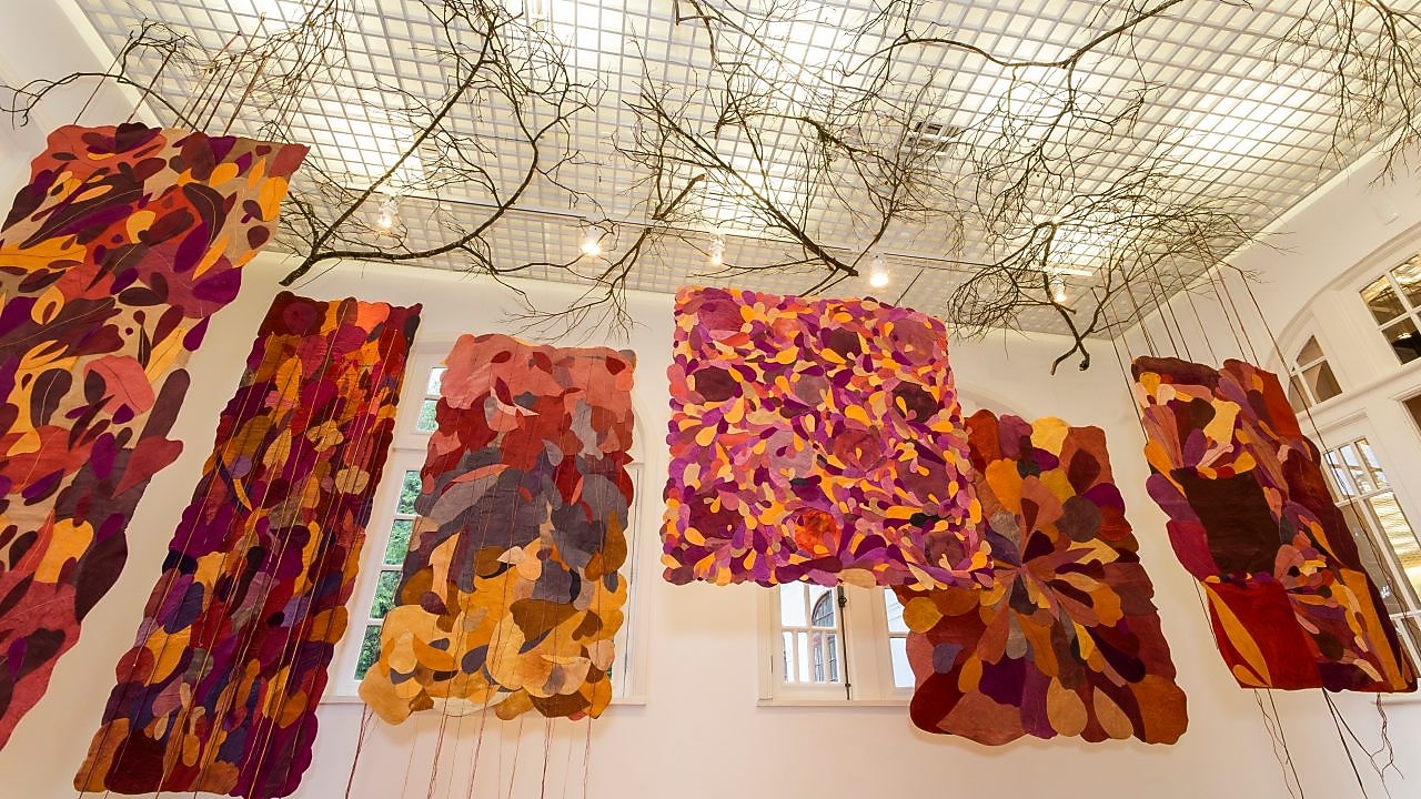  Instalação artística no Museu do Jardim Botânico do Rio de Janeiro com painéis têxteis suspensos em tons vibrantes e formas orgânicas. Galhos secos no teto complementam a atmosfera natural e imersiva.