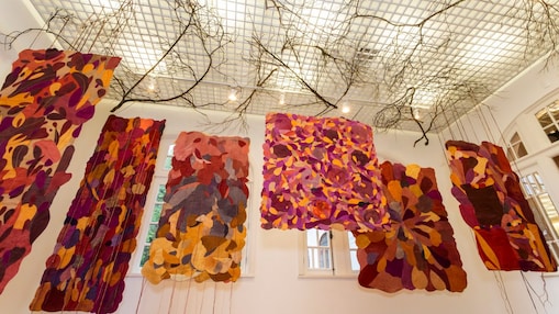  Instalação artística no Museu do Jardim Botânico do Rio de Janeiro com painéis têxteis suspensos em tons vibrantes e formas orgânicas. Galhos secos no teto complementam a atmosfera natural e imersiva.