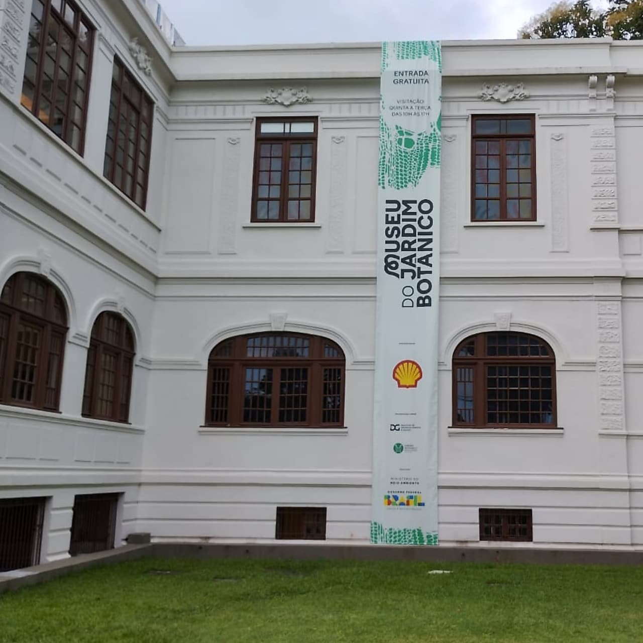 Fachada branca do Museu do Jardim Botânico, com janelas de moldura marrom e faixa vertical exibindo o nome do museu e logotipos de parceiros como Shell, sobre gramado verde em primeiro plano. 