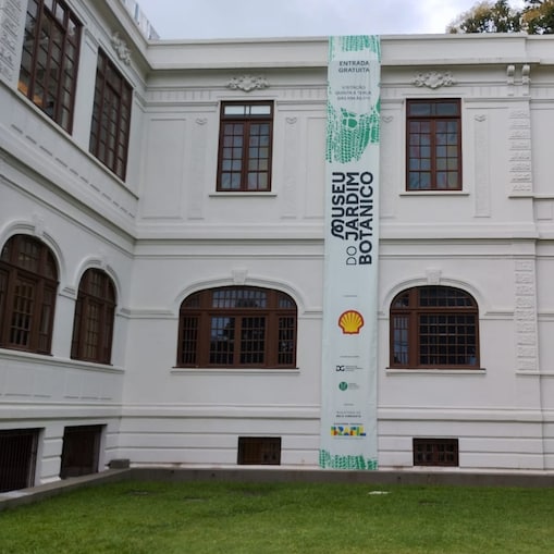 Fachada branca do Museu do Jardim Botânico, com janelas de moldura marrom e faixa vertical exibindo o nome do museu e logotipos de parceiros como Shell, sobre gramado verde em primeiro plano. 