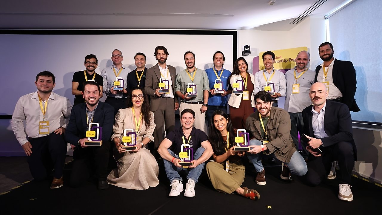 Grupo de pessoas sorrindo e posando em um auditório com troféus nas mãos durante um evento do programa Shell StartUp Engine. Todos usam crachás amarelos e demonstram celebração e reconhecimento.