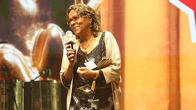 Pessoa em um palco segurando um microfone com a marca Shell e um troféu, enquanto fala. Usa vestido escuro com detalhes e um casaco claro, além de óculos. Ao fundo, há iluminação em tons quentes e elementos de cenário.