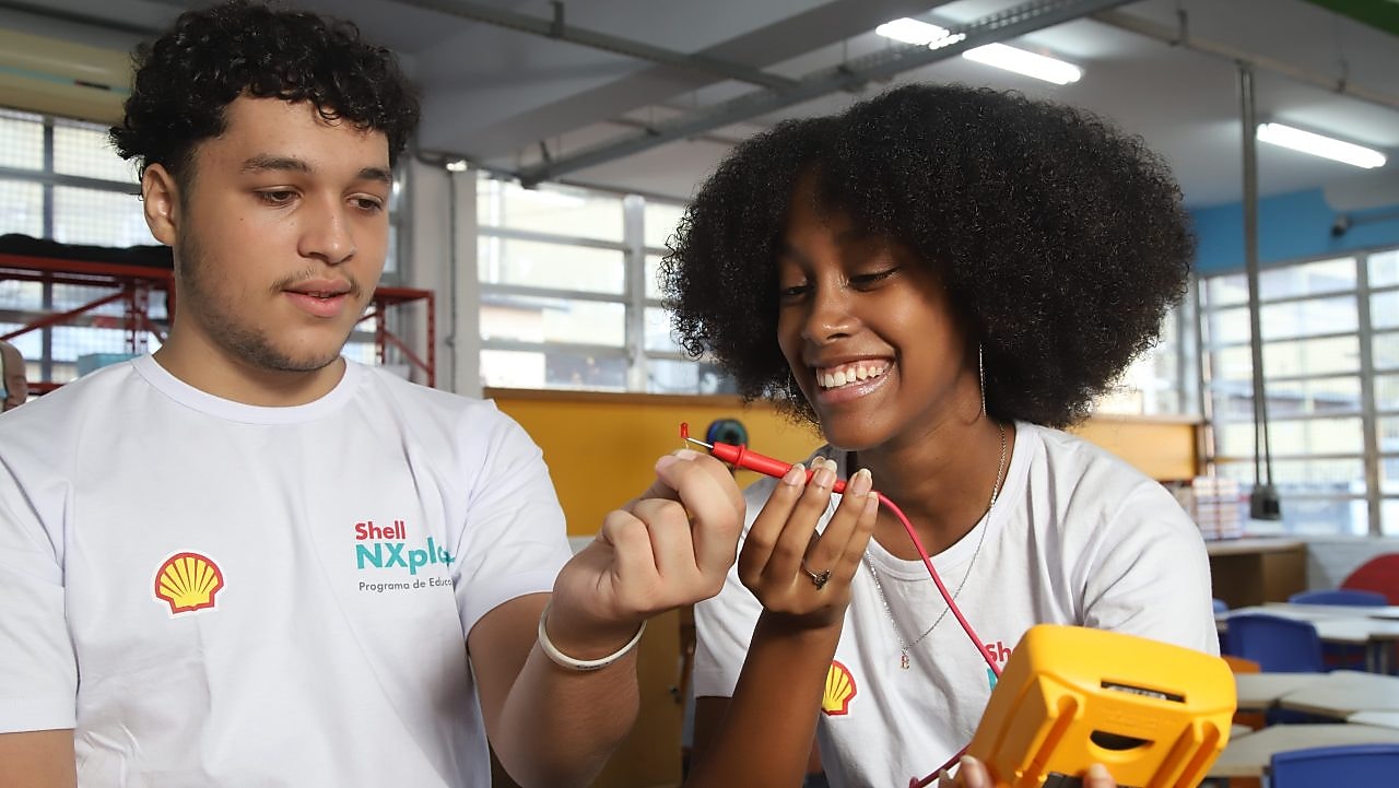 Dois jovens com camisetas do programa Shell NXplorers sorriem enquanto realizam uma atividade prática em sala de aula, manuseando fios e um equipamento eletrônico.