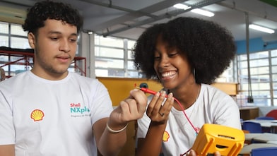 Dois jovens com camisetas do programa Shell NXplorers sorriem enquanto realizam uma atividade prática em sala de aula, manuseando fios e um equipamento eletrônico.