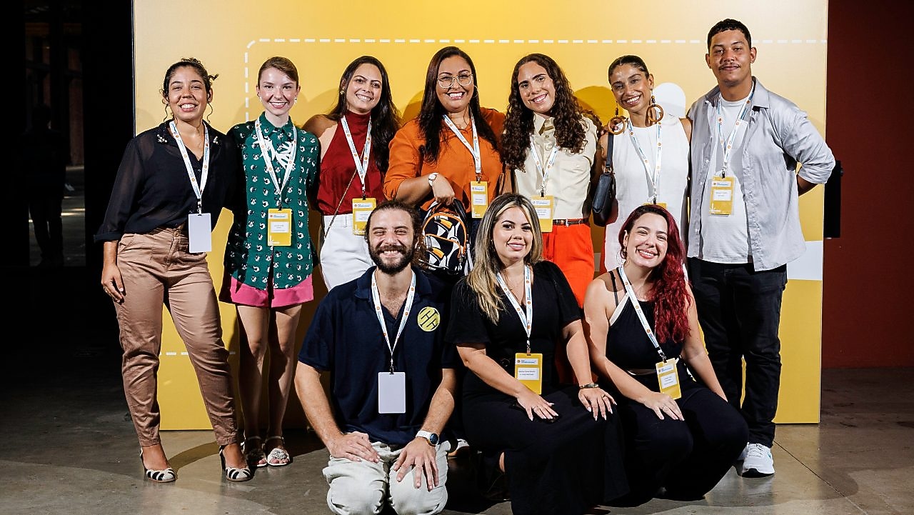 Um grupo de participantes sorrindo e posando juntos em frente a um painel amarelo do Shell Iniciativa Jovem durante um evento. Eles usam crachás de identificação e parecem fazer parte de um encontro profissional ou de empreendedorismo.