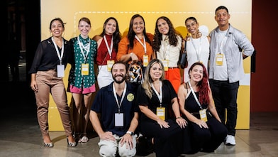 Um grupo de participantes sorrindo e posando juntos em frente a um painel amarelo do Shell Iniciativa Jovem durante um evento. Eles usam crachás de identificação e parecem fazer parte de um encontro profissional ou de empreendedorismo.