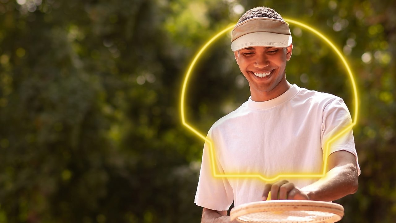 Jovem vestindo macacão de piloto azul sorri segurando um capacete amarelo com o logo da Shell Eco-marathon Brasil, sob um céu parcialmente nublado.