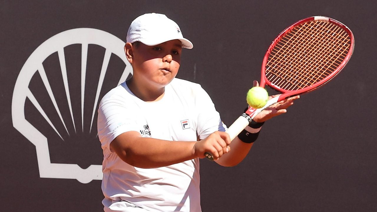 Jovem vestindo macacão de piloto azul sorri segurando um capacete amarelo com o logo da Shell Eco-marathon Brasil, sob um céu parcialmente nublado.