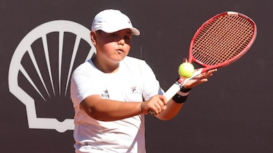 Jovem vestindo macacão de piloto azul sorri segurando um capacete amarelo com o logo da Shell Eco-marathon Brasil, sob um céu parcialmente nublado.