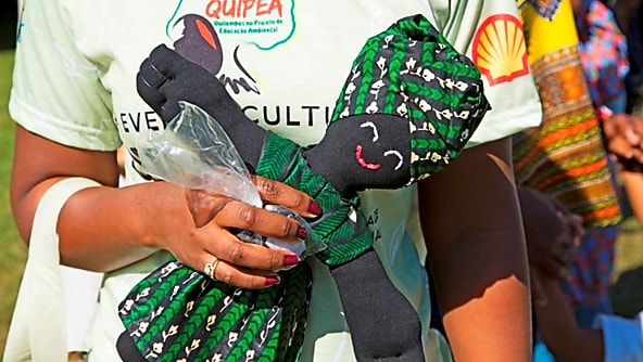 Pessoa vestindo camisa do projeto Quipea segura boneca de pano com roupas verdes estampadas durante evento cultural ao ar livre.